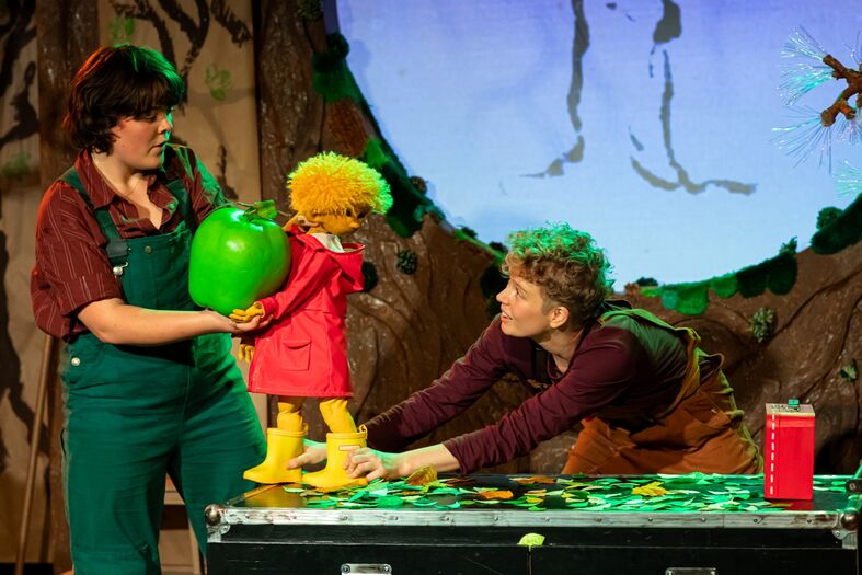 Twee acteurs op het podium met een groene appel en een pop in een kleurrijke, theatrale setting.