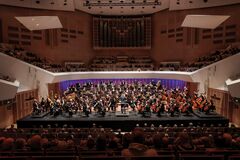 Orkest op het podium in een concertzaal, met een dirigent voor een groot publiek.