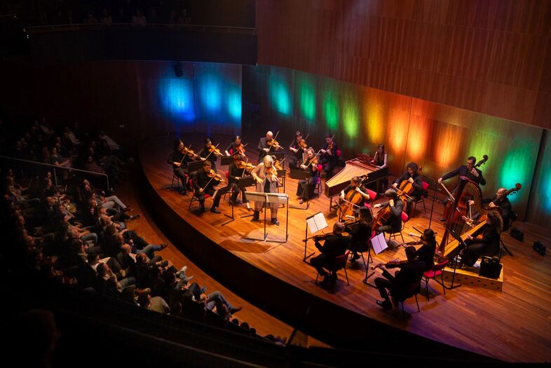 Een orkest speelt op een podium met gekleurde verlichting, terwijl het publiek erachter zit.