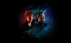 Two personen in donker geklede outfits staan tegen een vervallen gang, met beide gezichten in verschillend gekleurd licht en silhouetten in de achtergrond.