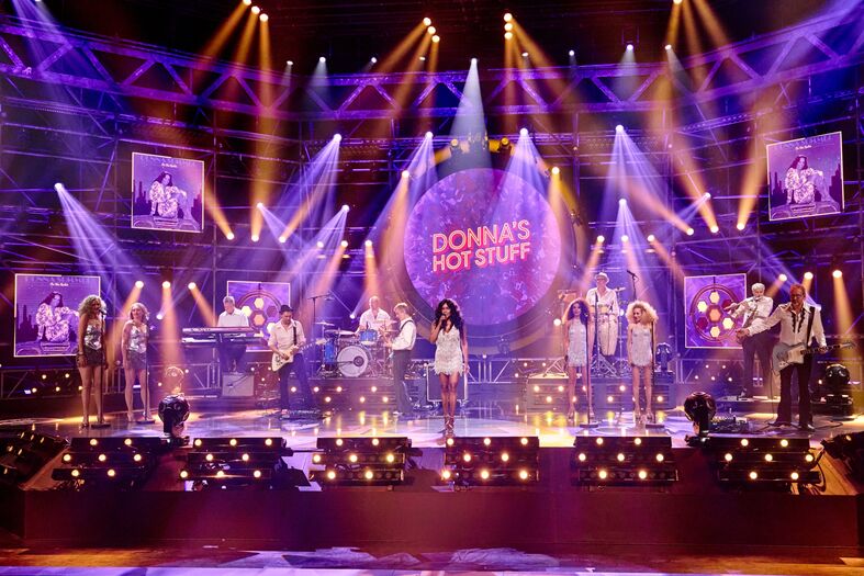 Grote band met zanger en dansers optredend onder felgekleurde podiumlichten.