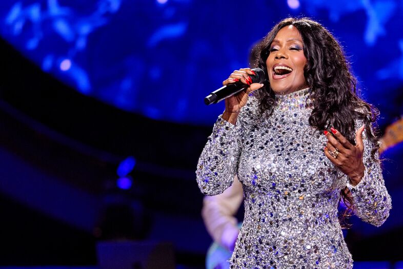 Persoon in glinsterende outfit zingt op een felverlicht podium met microfoon.