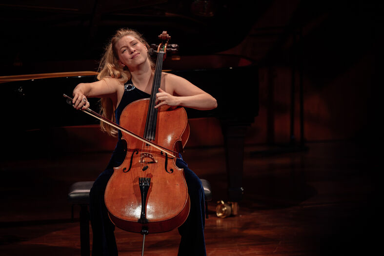 Een vrouwelijke celliste speelt emotioneel op een podium, zittend op een stoel met een cello.