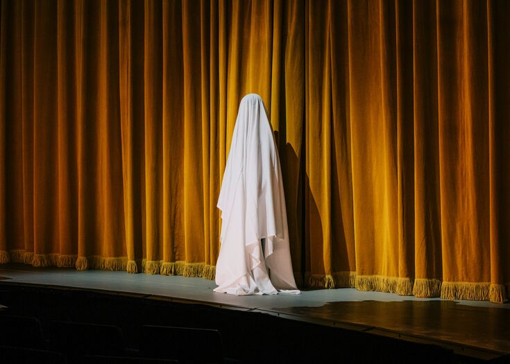 Een spookachtige figuur in een witte doek staat achter een gouden theatergordijn.