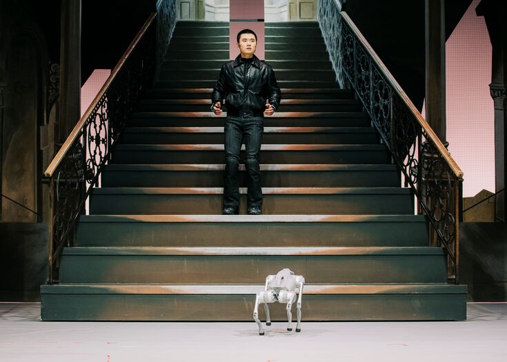 Een man in een zwart leren jack staat op de trap met een klein robotachtig apparaat voor hem.