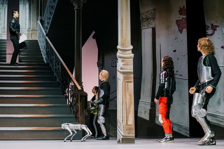 Een podium met acteurs in futuristische outfits, een hond en een trap op de achtergrond.