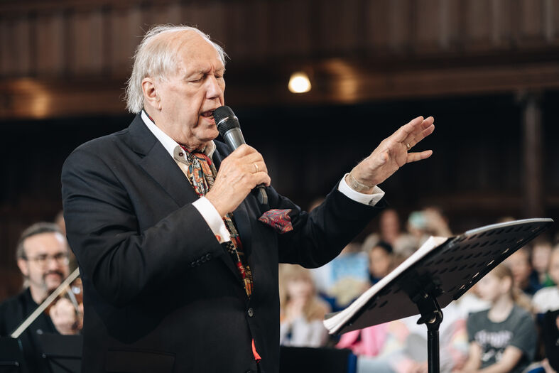 Een oudere man in een zwart vest houdt een microfoon vast en dirigeert een koor in een zaal.