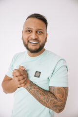 Man met een tattoo op zijn rechterarm, glimlachend en in een mintgroen T-shirt, staat met gekruiste handen voor een witte achtergrond.