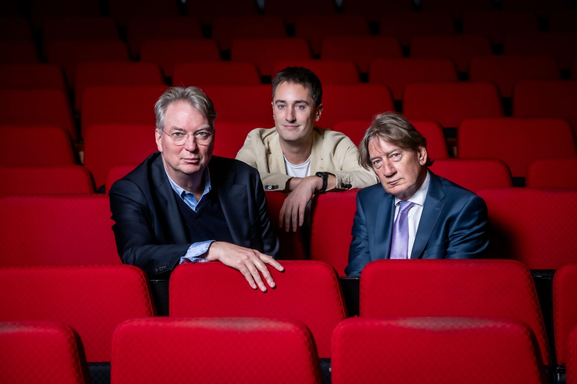 Drie mannen zitten op een rode theaterstoel, met gefocuste blikken, in een lege zaal.