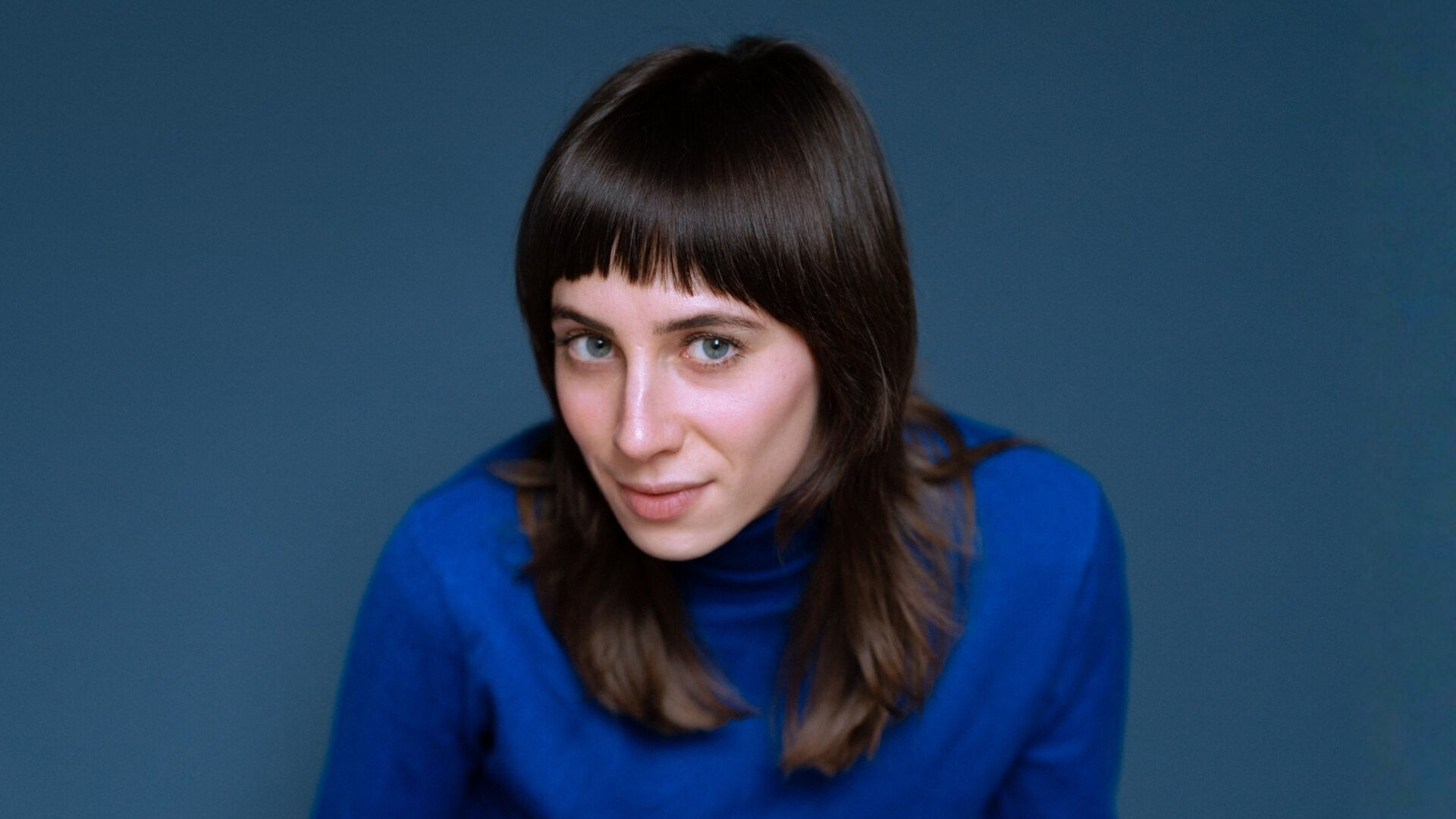 Een vrouw met lang, bruin haar en een rechte pony, gekleed in een blauwe trui, kijkt nieuwsgierig in de camera.