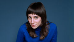 Een vrouw met lang, bruin haar en een rechte pony, gekleed in een blauwe trui, kijkt nieuwsgierig in de camera.