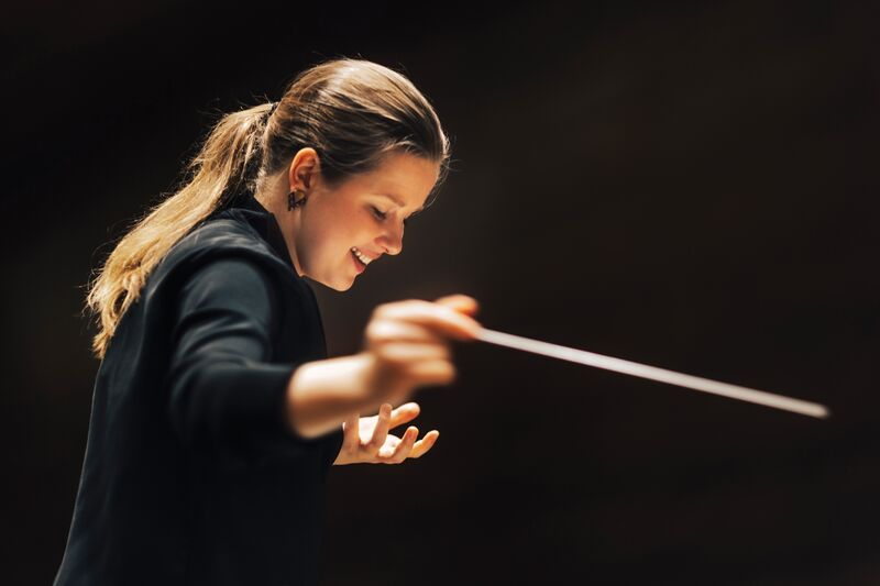 Persoon dirigeert met baton in warme spotlight tegen donkere lege achtergrond.