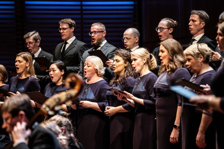 Een koor van zangers in donkere kleding, dat samen zingt op een podium met een muzikant op de voorgrond.