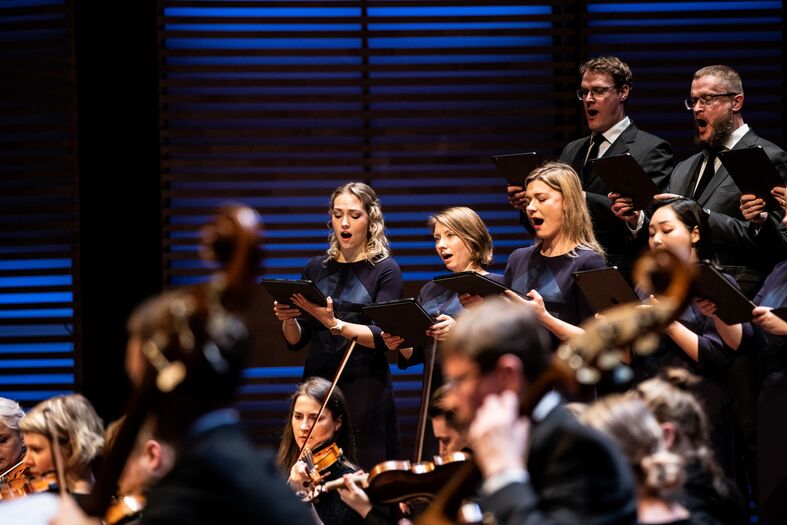 Een koor van zangers in formele kleding voert muziek uit op een podium met een orkest.