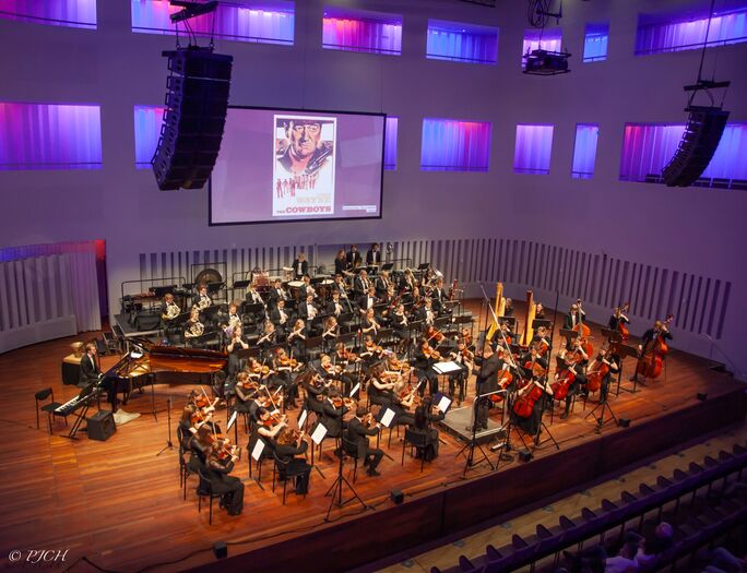 Een orkest speelt op een podium in een concertzaal met kleurrijke verlichting en een groot scherm op de achtergrond.