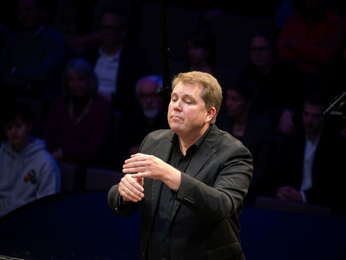 Een dirigent leidt een orkest op het podium, met publiek op de achtergrond in een dimlicht.