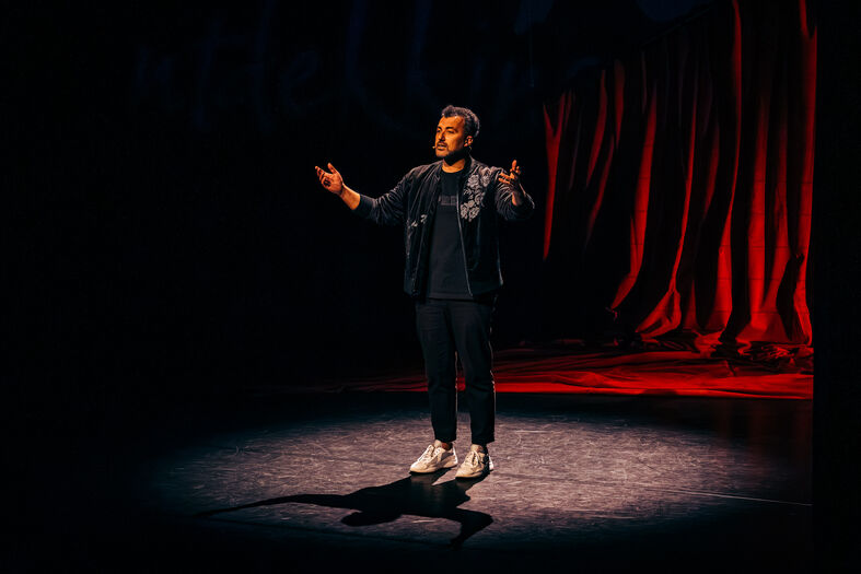 Man staat op een podium in de schijnwerpers, met zijn handen omhoog, voor een rode achtergrond.