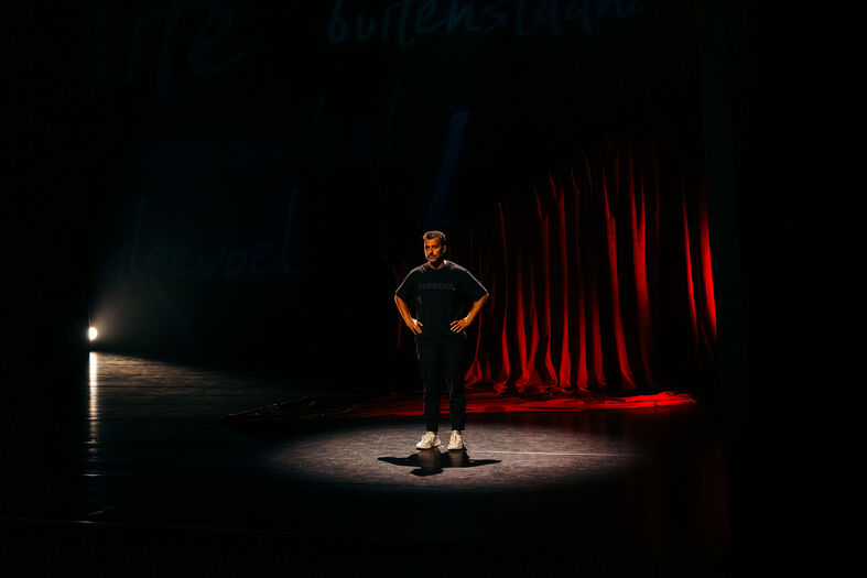 Een performer staat in spotlicht op een podium met rode gordijnen op de achtergrond.