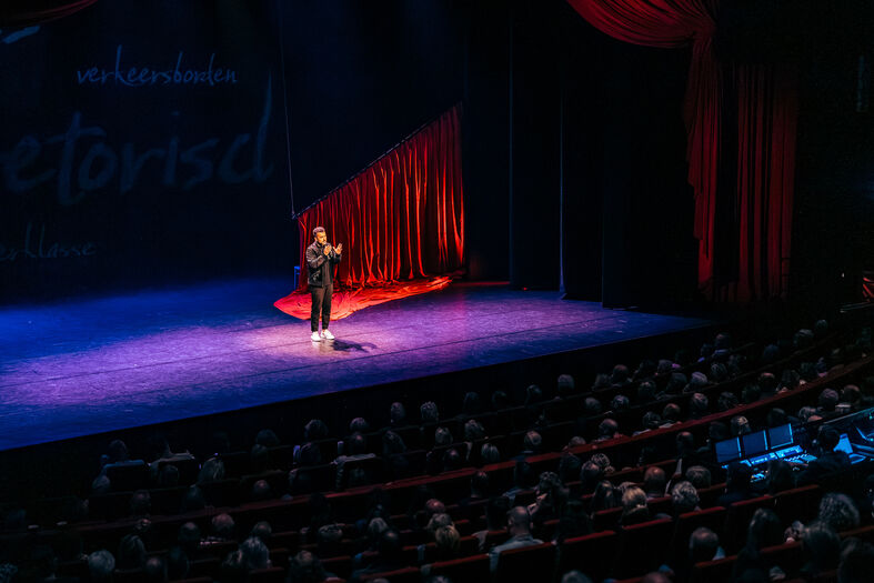 Een performer staat op het podium van een theater voor een publiek, met kleurrijke gordijnen op de achtergrond.