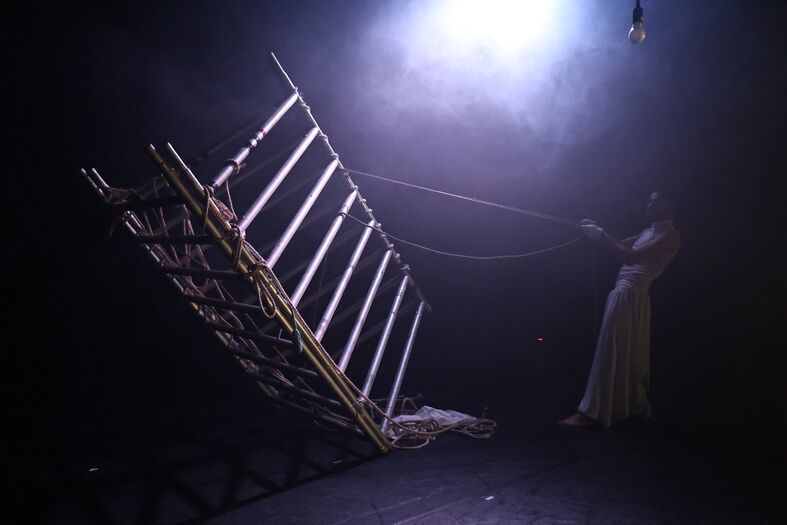 Een vrouw in een lange jurk trekt aan een touw waaraan een hellende houten constructie hangt, omgeven door mist en licht.