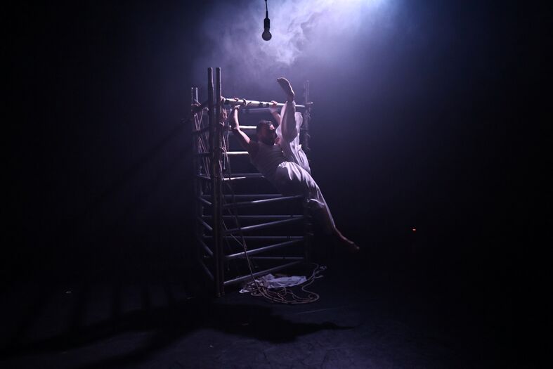 Een persoon in witte kleding zit op een ladder in een donker, mistig ruimtescenario met een enkele lichtstraal.