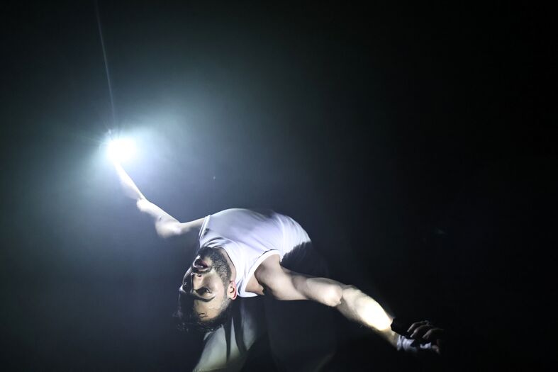 Een performer in een witte top maakt een acrobatische beweging met een lichtbundel in zijn hand in een donkere omgeving.