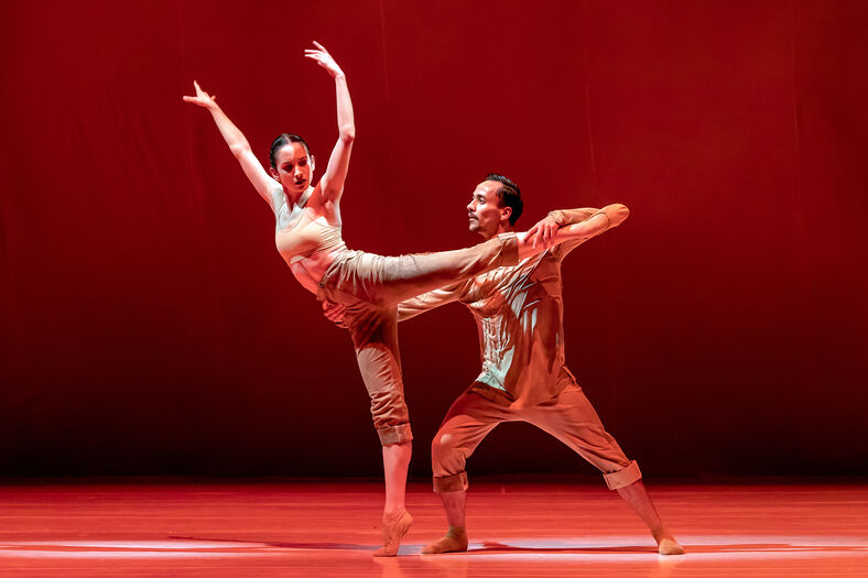 Twee dansers op een rood verlicht podium, waarbij de ene een been hoog optilt terwijl de partner steun biedt.