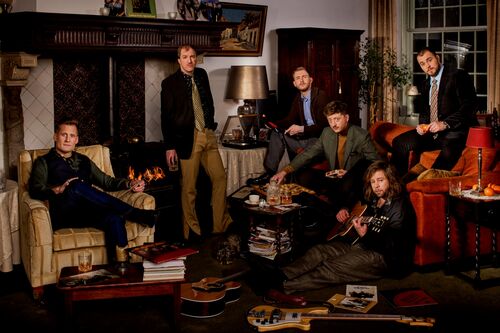 Een groep van zes mannen in formele kleding poseert in een sfeervol ingerichte woonkamer.