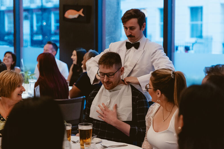 Een ober kleedt een man in een restaurant aan met een servet, terwijl gasten aan tafel glimlachen en met elkaar praten.