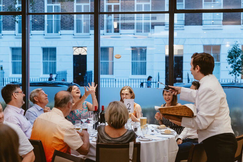 Een ober serveert een gerecht aan een groep mensen aan een ronde tafel in een restaurant met grote ramen.