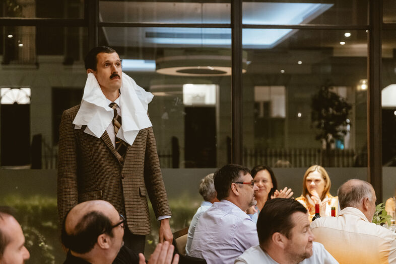 Man in een bruine blazer met een servet om zijn nek, staat op tijdens een diner terwijl gasten applaudisseren.