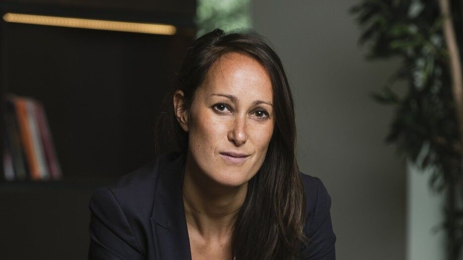 Zakelijke portretfoto van een vrouw in een zwart pak, zittend op een rode stoel met een neutrale achtergrond.