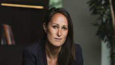 Zakelijke portretfoto van een vrouw in een zwart pak, zittend op een rode stoel met een neutrale achtergrond.
