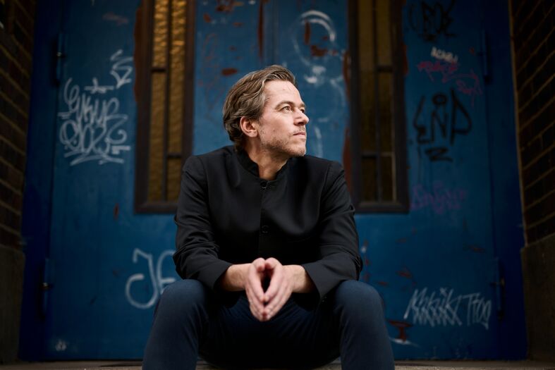 Man met een contemplatieve uitdrukking zit op de grond voor een met graffiti bedekte blauwe muur.