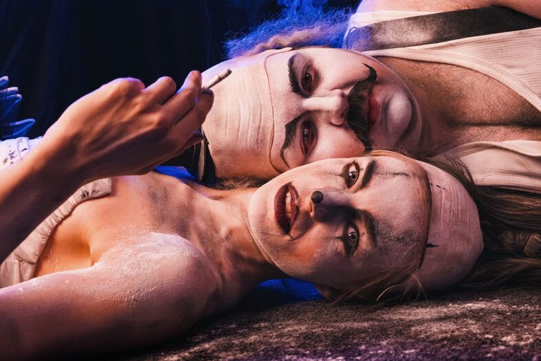 Twee mensen in clownkostuums met witte make-up liggen op de grond, met een dramatische en expressieve uitstraling.