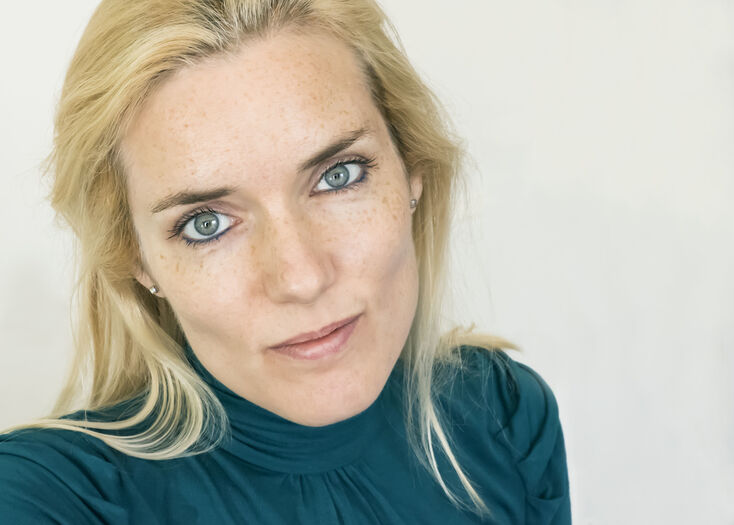 Portret van een vrouw met blonde haren en blauwe ogen, gekleed in een groene trui, met een neutrale achtergrond.
