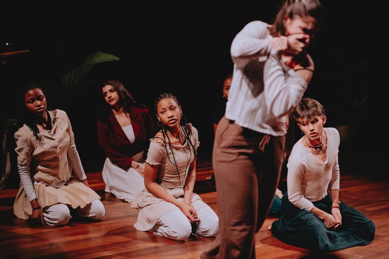 Groep performers knielt en zit op houten podiumvloer, terwijl één persoon vooraan bewegend onscherp in beeld is.