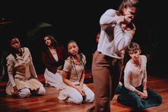 Groep performers knielt en zit op houten podiumvloer, terwijl één persoon vooraan bewegend onscherp in beeld is.