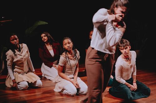 Groep performers knielt en zit op houten podiumvloer, terwijl één persoon vooraan bewegend onscherp in beeld is.