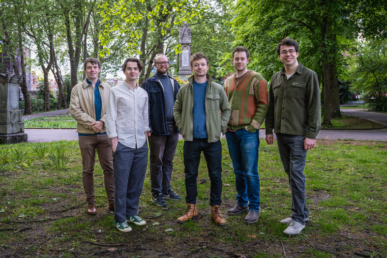 Zes mannen poseren in een park, omringd door bomen, en kijken naar de camera.
