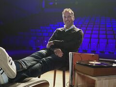 Man zit relaxed op een stoel met zijn voeten op een tafel, in een lege theaterzaal met blauwe verlichting.