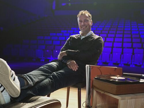 Man zit relaxed op een stoel met zijn voeten op een tafel, in een lege theaterzaal met blauwe verlichting.