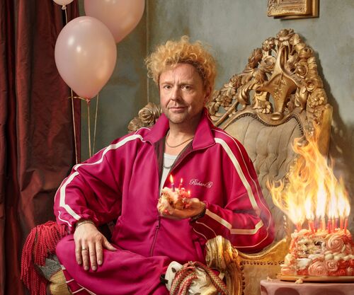 Richard Groenendijk in roze trainingspak zit op sierlijke stoel naast brandende taart en ballonnen