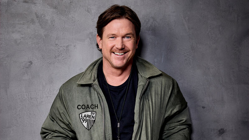 Lachende Johnny de Mol in groene coachjas staat ontspannen tegen een grijze muur met handen in de zakken.