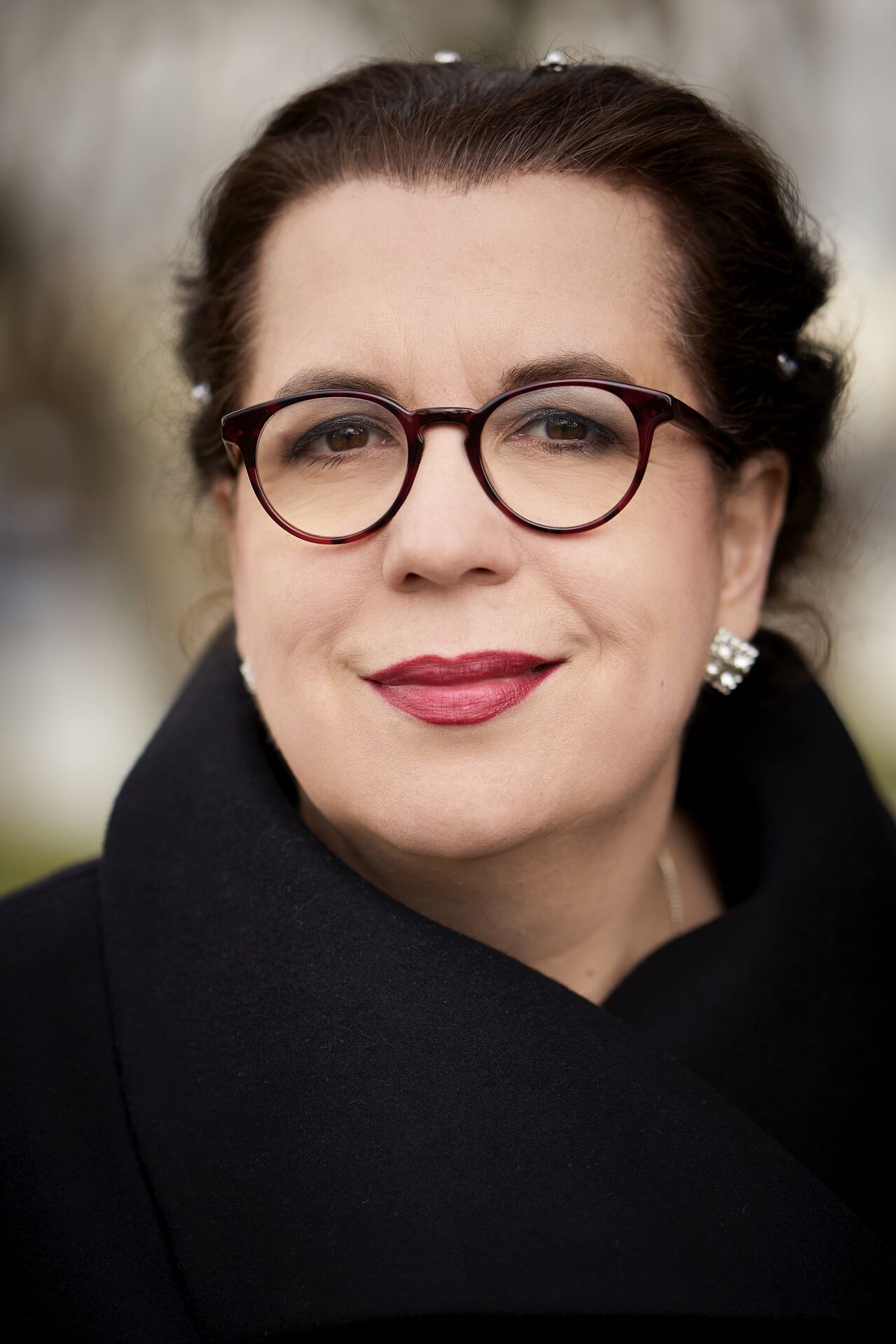 Professioneel portret van een vrouw met bruin haar, ronde bril en een zwarte jas, met een neutrale achtergrond.