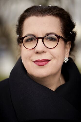 Professioneel portret van een vrouw met bruin haar, ronde bril en een zwarte jas, met een neutrale achtergrond.
