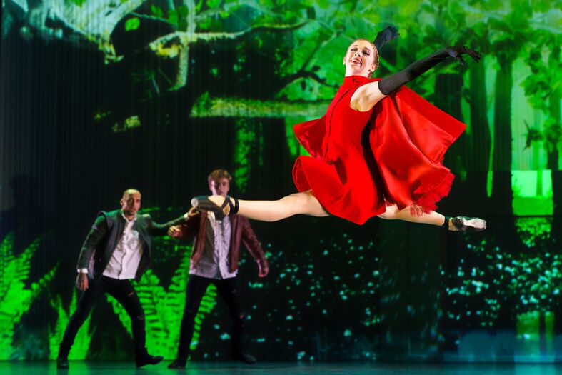 Danseres in rode jurk maakt een sprongetje, geflankeerd door twee performers op een groene achtergrond.