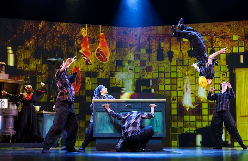 Vijf dansers in een theatrale setting, met één persoon in de lucht terwijl anderen reageren op een achtergrond van keukenitems.