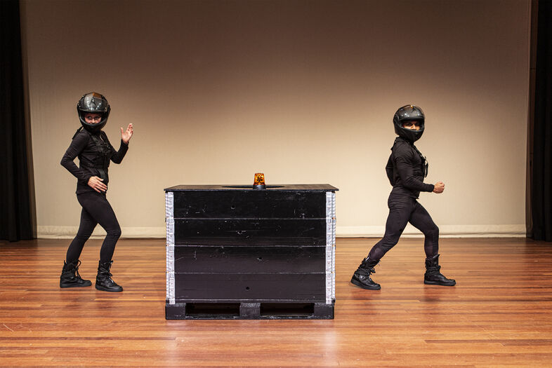 Twee dansers in zwarte outfits en helmen voeren een choreografie op rond een zwarte kist op een podium.
