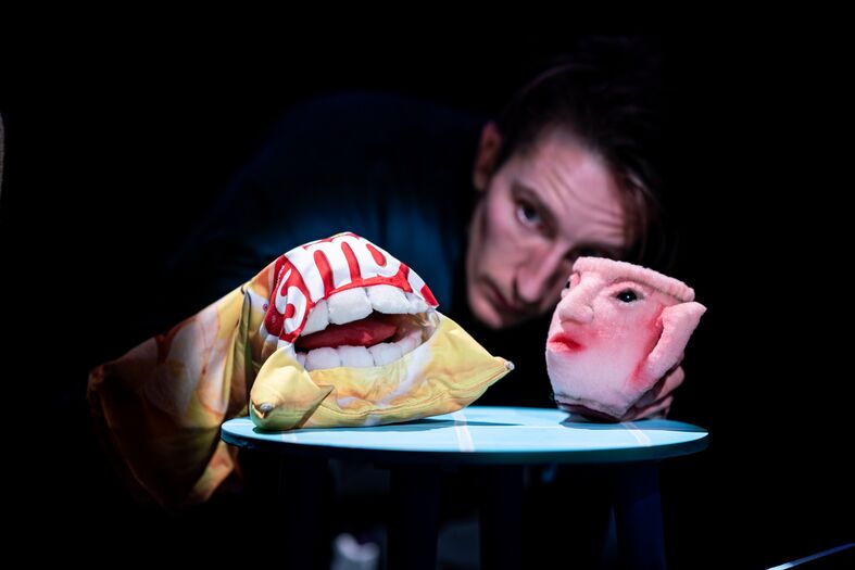 Performer met een pop in de vorm van een gezicht en een andere pop met een grote mond, die op een tafel is geplaatst.